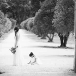 Photo en noir et blanc d'un portail ouvert sur une allée d'oliviers où se tiennent une mariée de dos et une enfant tenant son voile