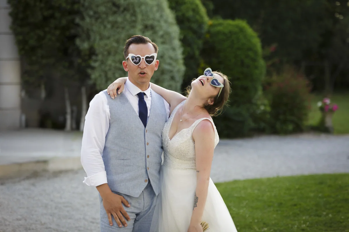 Photo d'un jeune couple de mariés avec leurs lunettes cœurs. La mariée le tient par le cou et penche sa tête en arrière dans une simulation d'épuisement
