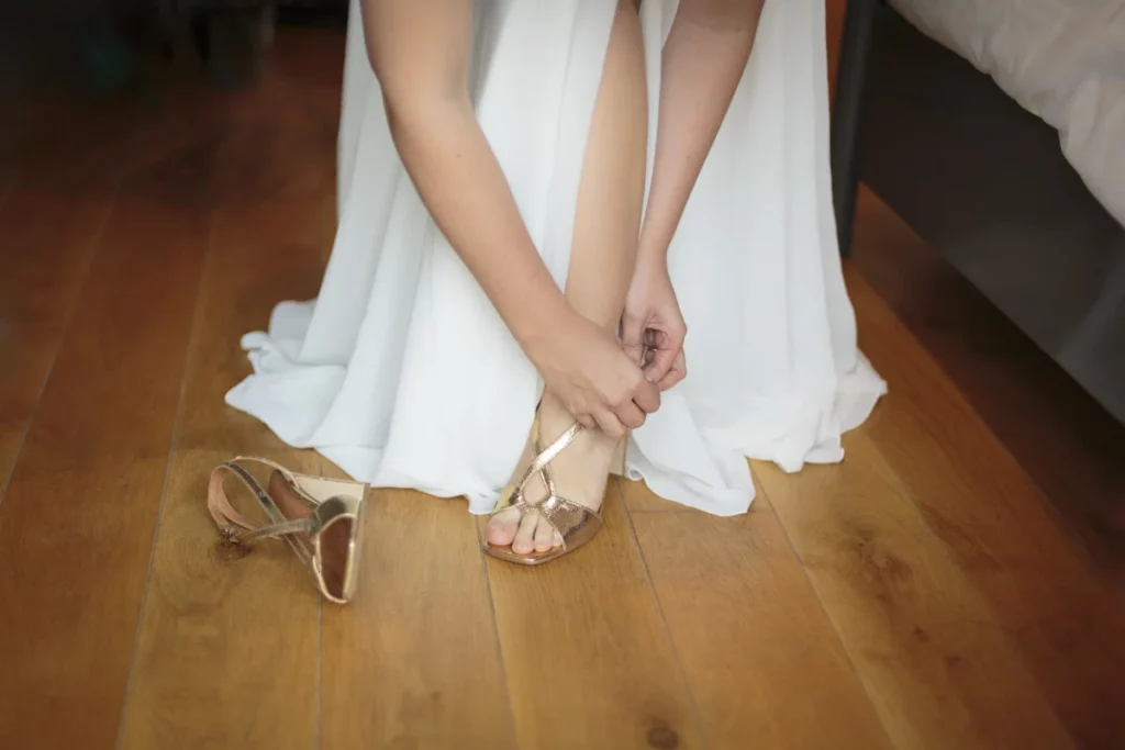Gros plan sur la mariée qui chausse ses sandales dorées. On ne voit que le bas de sa robe et ses avant-bras