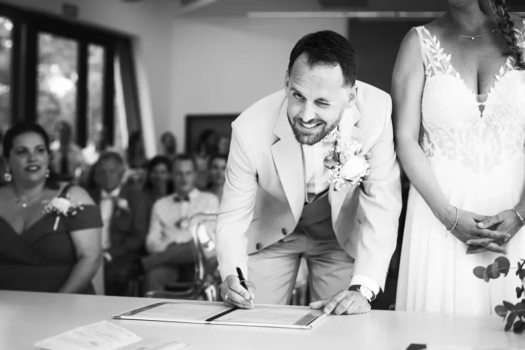 Photo en noir et blanc du marié faisant un clin d'œil au photographe pendant qu'il signe à la mairie