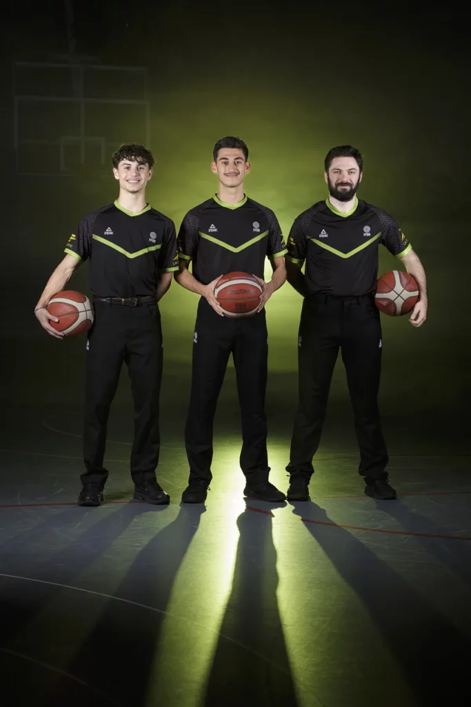 Portrait de trois arbitres de baskets tenant chacun un ballon, dans un fond noir brumeux éclairé de jaune à l'intérieur d'un gymnase