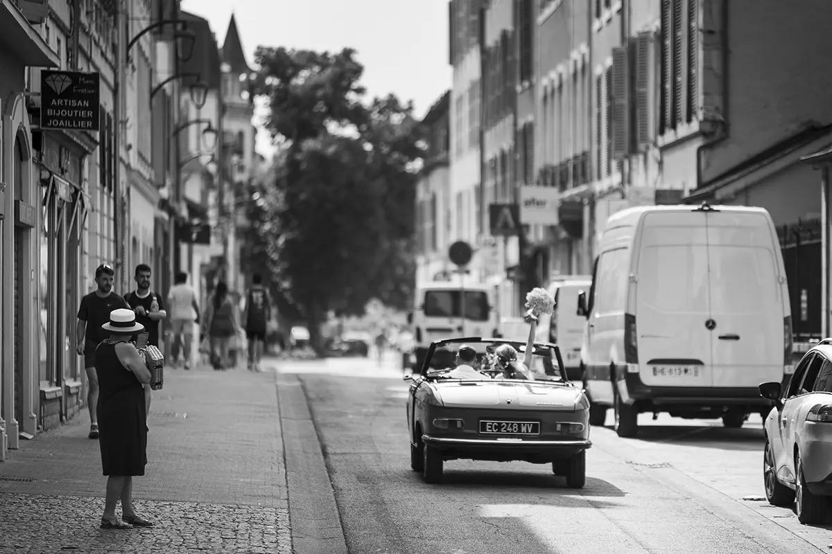 Photo en noir et blanc où l'on aperçoit les mariés dans une voiture rétro décapotable de dos roulant dans une rue de Pau