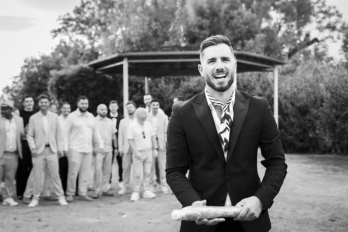 Photo en noir et blanc d'un marié s'apprêtant à lancé un saucisson aux hommes d'une assemblée derrière lui comme s'il lançait un bouquet