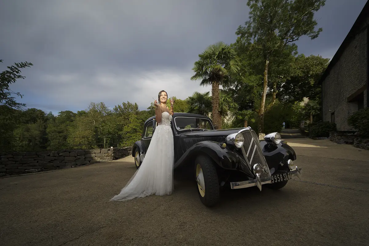 Photo d'une mariée accoudée à un voiture vintage, tenant une coupe de champagne dans une main et une cigarette dans l'autre