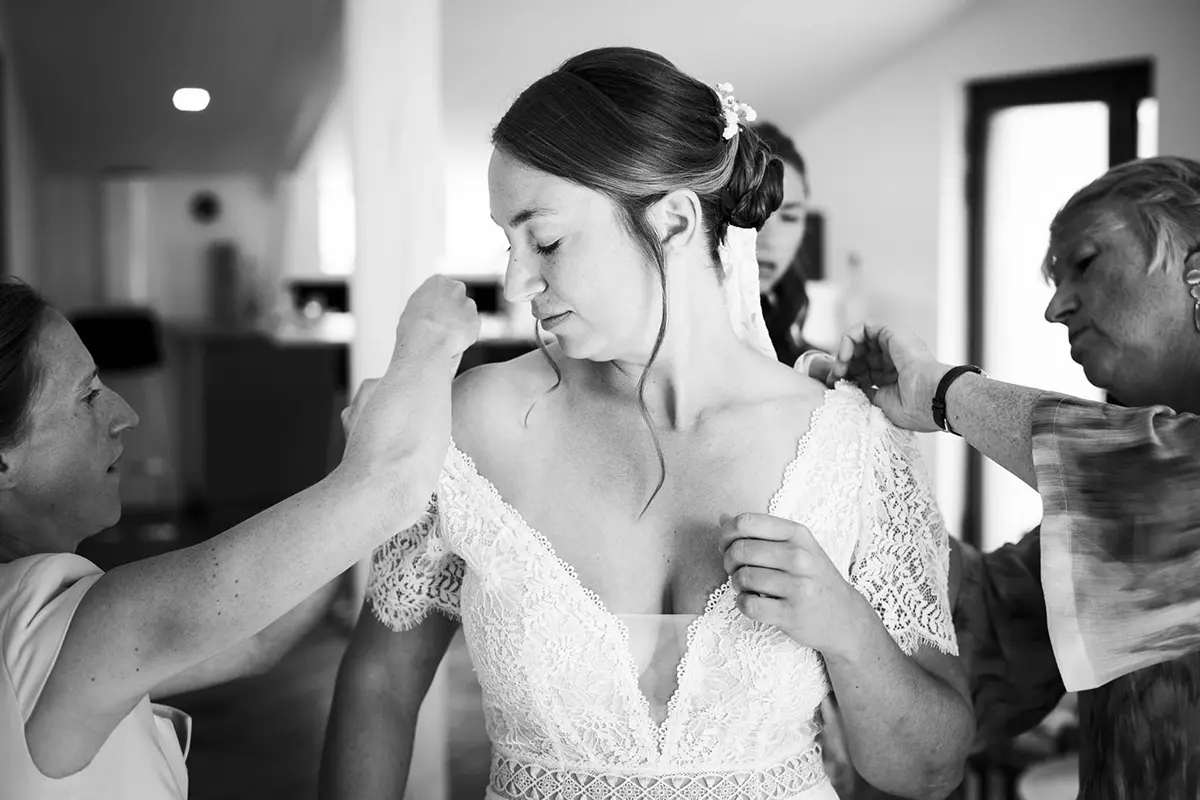 Photo en noir et blanc d'une mariée en pleine préparation, deux femmes s'affairent autour d'elle pour ajuster sa robe aux épaules