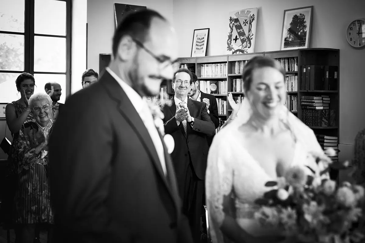 Photo en noir en blanc prise lors de la cérémonie d'un mariage à la mairie. La mise au point est faite sur un invité heureux dans l'assemblée. On peut l'apercevoir entre les deux mariés qui sont au premier plan