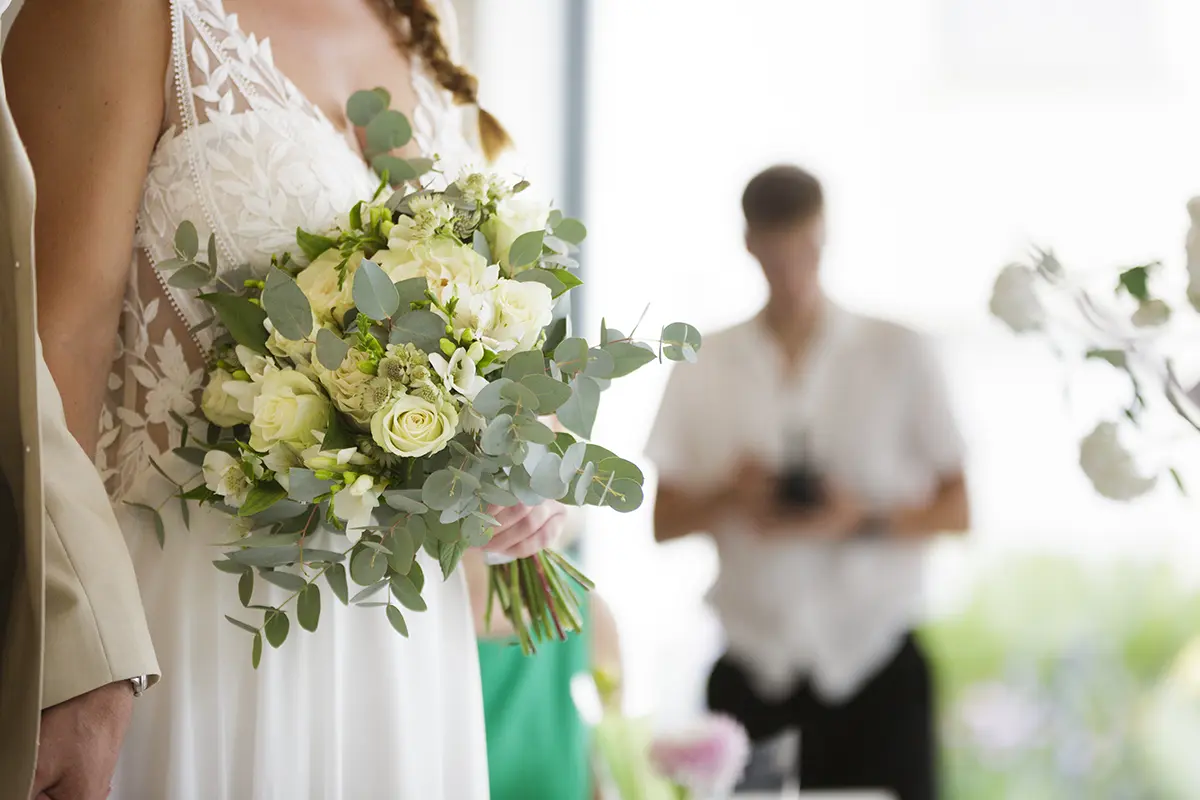 Gros plan sur le bouquet de la mariée, on distingue au loin une personne la prenant aussi en photo