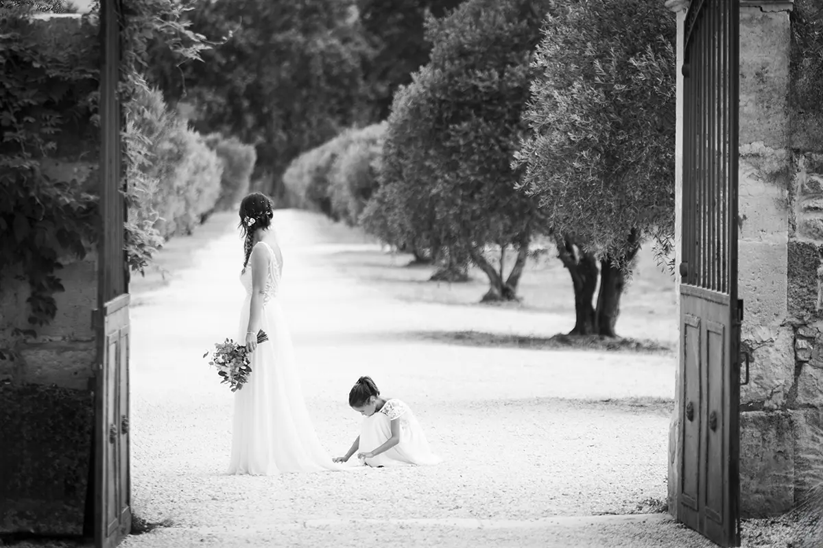 Photo en noir et blanc d'une mariée devant l'entrée d'un domaine ou elle regarde l'allée d'oliviers avec une petite fille qui ajuste sa robe en bas